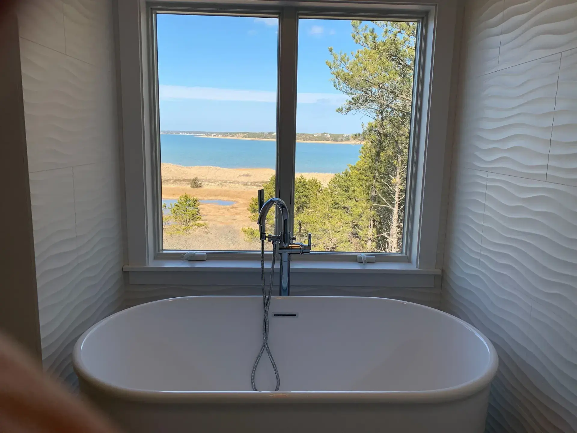 Freestanding soaking tub beneath a window overlooking Cape Cod marsh and water by Tuleika Building Company